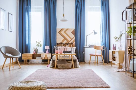 freundlich eingerichtetes Schlafzimmer mit Bett, Teppich, Dekoration und blauen Gardinen an zwei Fenstern